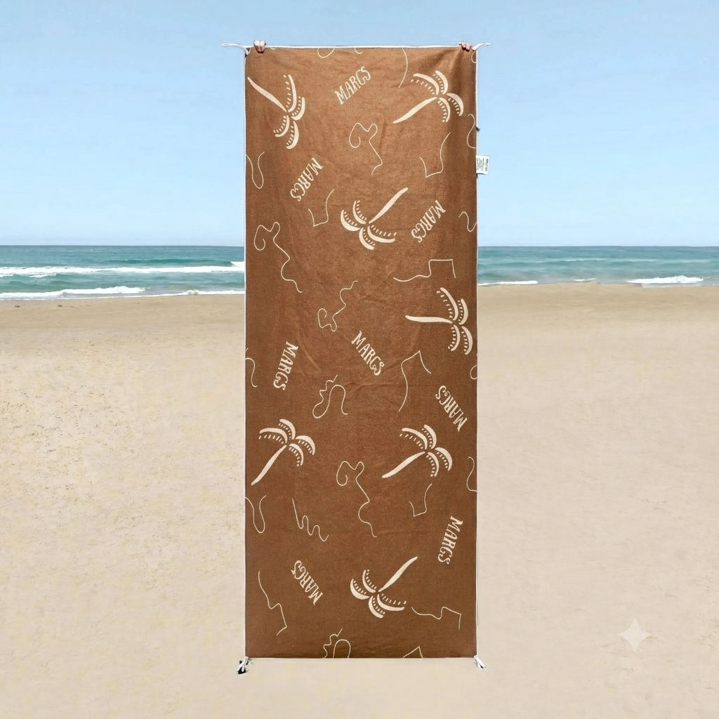 A brown beach towel with white text and designs, displayed on a beach setting.
