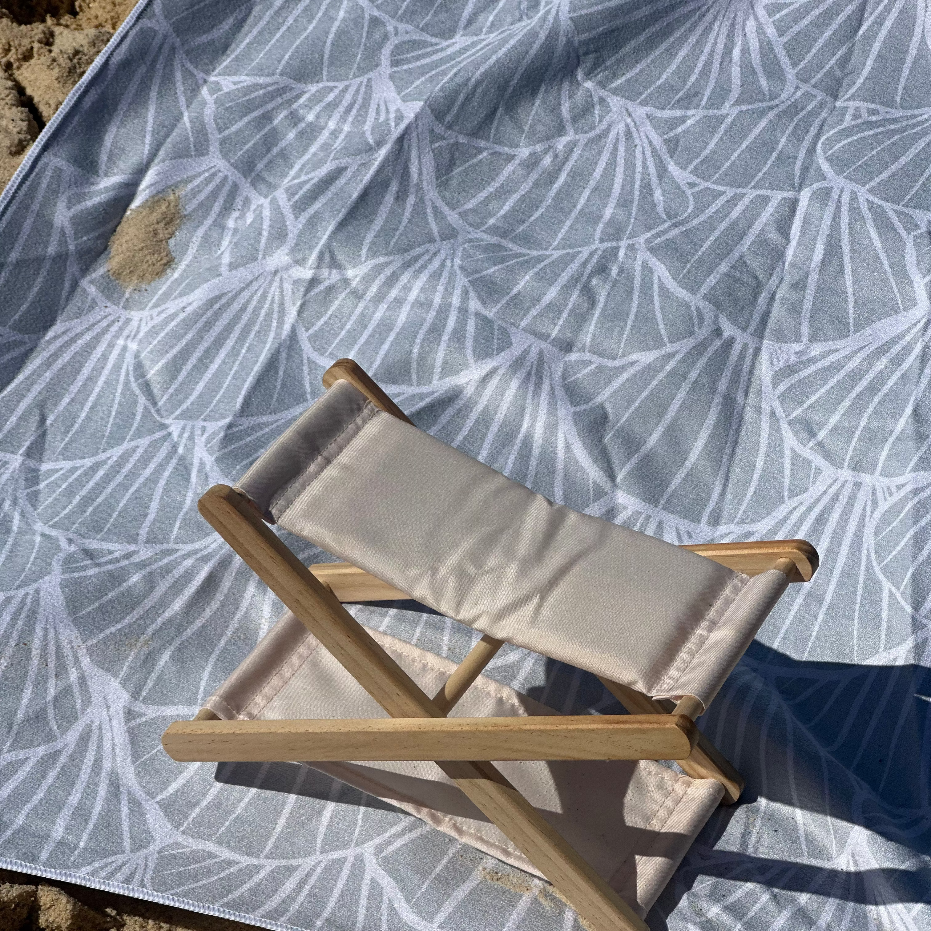 A beach headrest placed on a sand-free beach towel with a soft shell print design, creating a comfortable and stylish spot to relax by the sea.