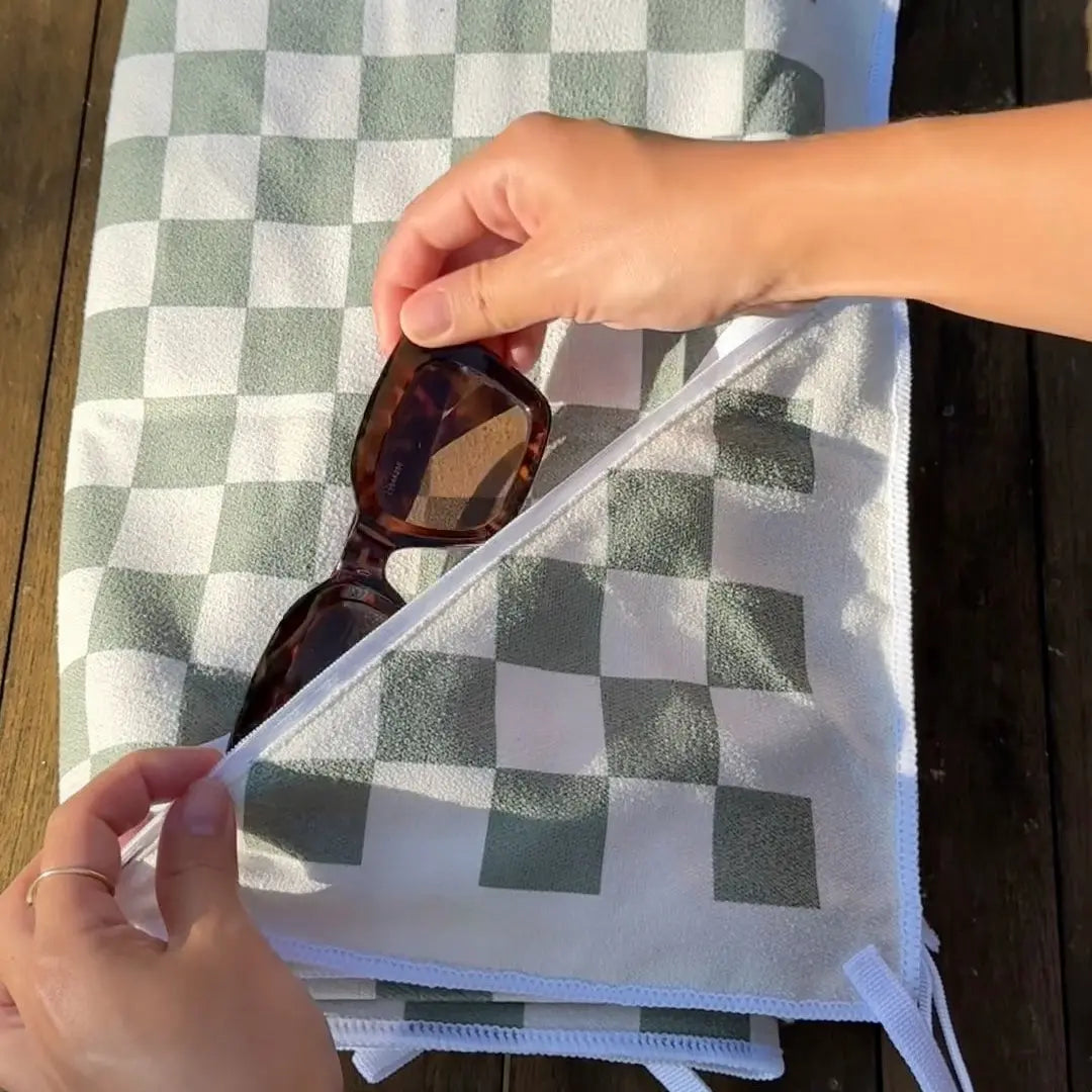 Person holding a green checkered sand free beach towel and putting sunglasses in the zip pocket on a wooden surface