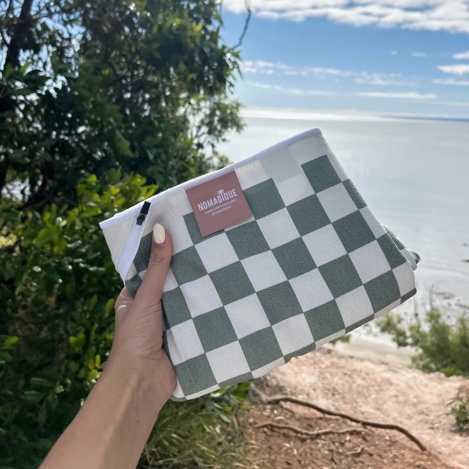 green Checkered beach towel with pocket held by a hand with a scenic background of trees and water.