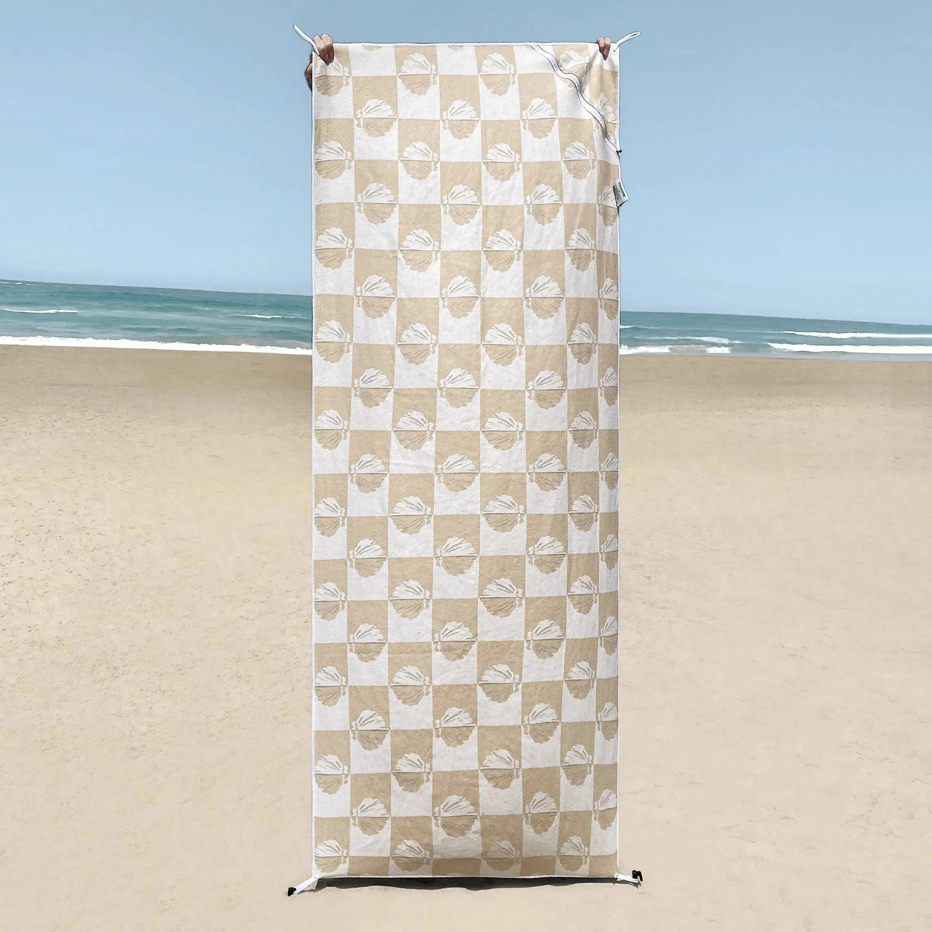 A beige and white beach towel with a shell pattern is displayed upright on a sandy beach, with the ocean and sky in the background.