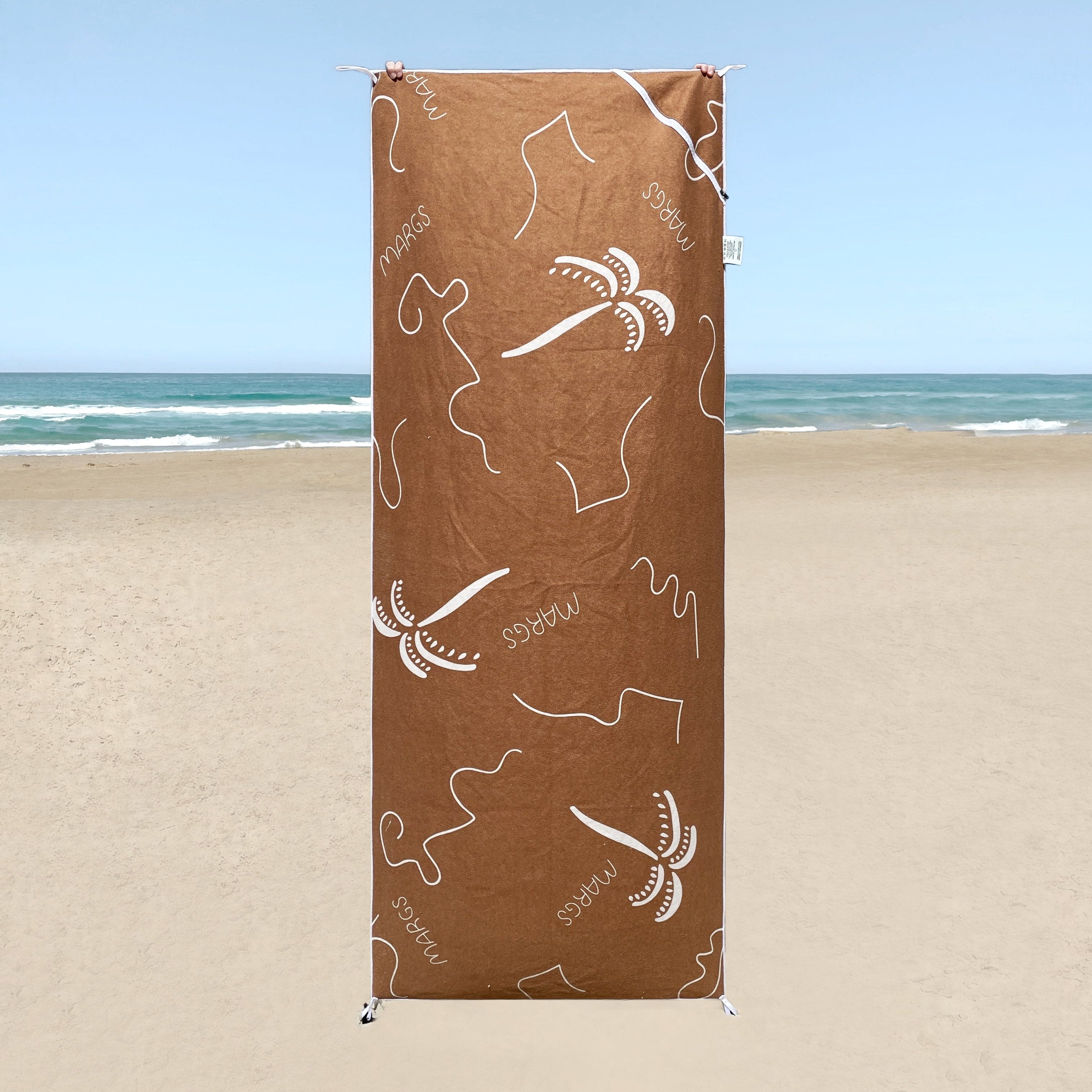 A brown beach towel with white text and designs, displayed on a beach setting.