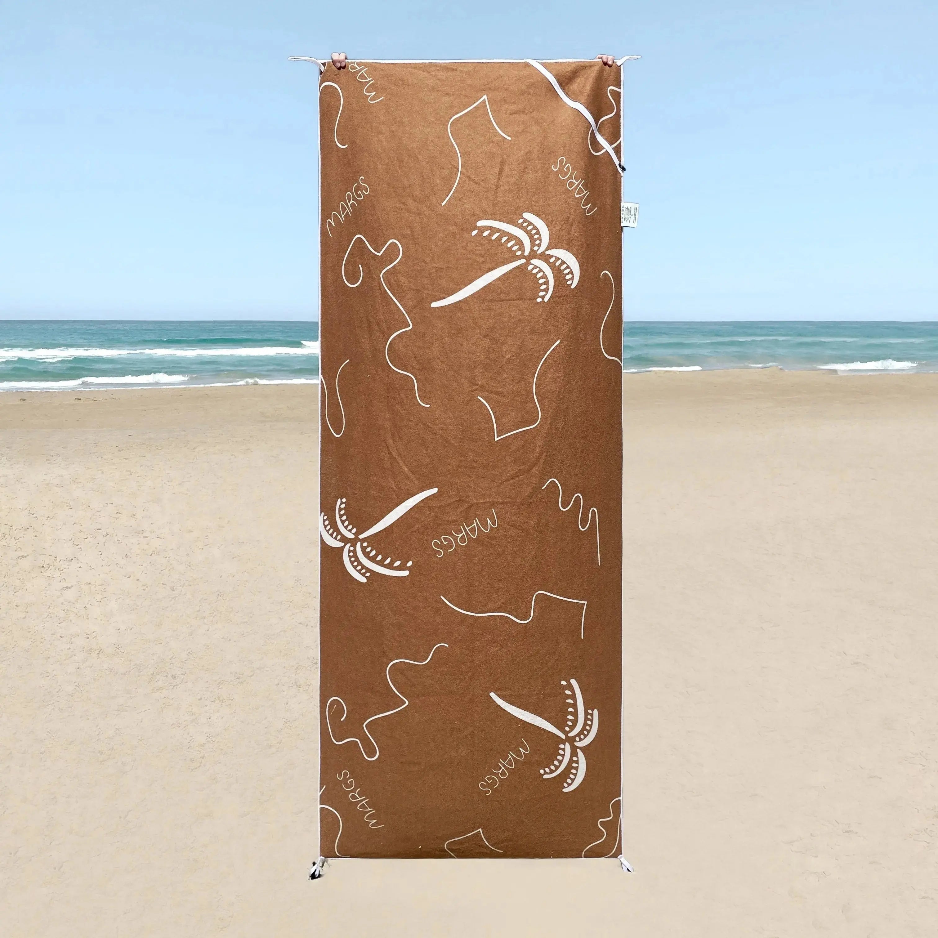 A brown beach towel with white text and designs, displayed on a beach setting.