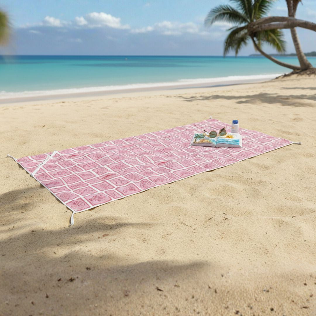 The towel is finished with a lighter-coloured pink border that frames the pattern neatly, helping define its shape and separate it from the surrounding sand.