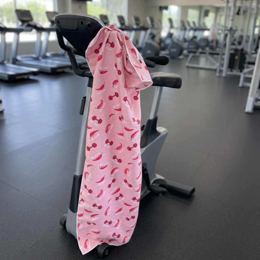 A bright pink towel with a quirky chili-and-dumbbell print hangs over a workout equipment bringing humor and attitude into a clean, minimalist gym setting.