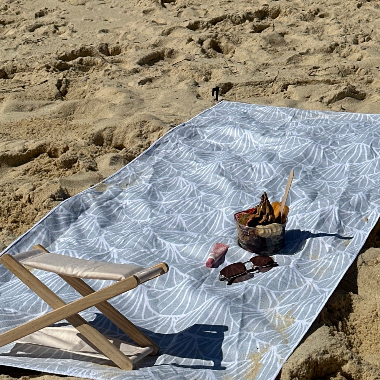 A sand-free beach towel with a soft shell print is spread across the sandy beach, creating a clean and comfortable lounging space. A wooden beach headrest sits at one end of the towel for support while relaxing. On the towel are a pair of sunglasses, a tube of lip balm, and a bowl of snacks topped with fruit and chocolate, capturing the perfect laid-back beach moment under the sun.