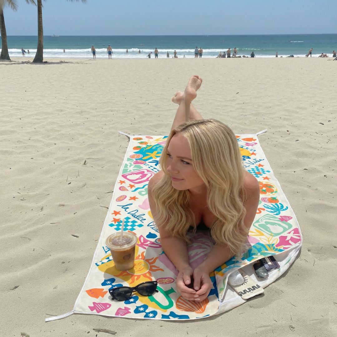 Woman sitting on a colourful beach towel with a drink and sunglasses at the beach.